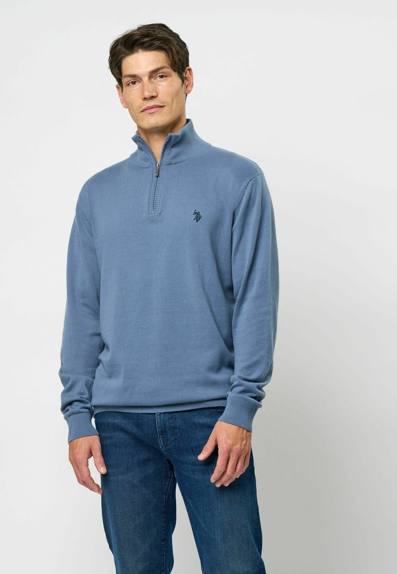 Kék pulóver, negyedcipzáros gallérral, bordás mandzsettákkal és szegéllyel. A mellkasán egy kis fekete logó található. Puha anyagból készült.