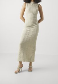 Knee-length, bez rukávů, světle béžové šaty s texturovaným povrchem; doplněné šedými sandály na podpatku. Jednoduchý design, přiléhavá silueta.