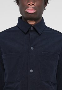 Camisa de pana azul marino con cuello, que cuenta con dos bolsillos delanteros y cierre de botones, tejido texturizado con estrías verticales.