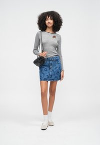 Szürke hosszú ujjú felső barna kör alakú díszítéssel, kék virágmintás farmer szoknyával párosítva. A modell fehér zoknit és világos cipőt visel.