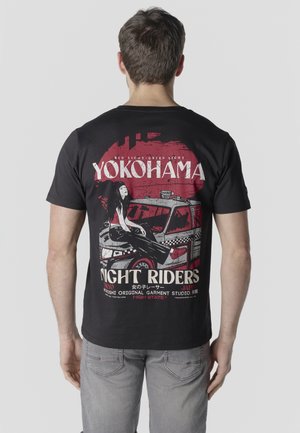 Hombre con camiseta negra que lleva un gráfico rojo y blanco de una mujer sentada en un taxi y texto que dice "Yokohama Night Riders Tokyo Japan".