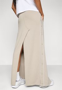 Beige maxi skirt in soft fabric, featuring a side slit, vertical button detail, and white piping along the seam. Pockets included.