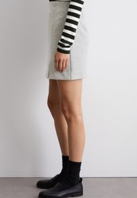 Grauer Rock mit einem seitlichen Schlitz, kombiniert mit einem schwarz-weißen gestreiften Langarmoberteil. Die Fußbekleidung umfasst schwarze Slipper und dunkle Socken.