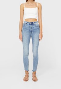 Mujer vestida con jeans ajustados de talle alto en azul claro con ligeras desgastaciones, un top corto blanco y sandalias planas de tiras en tono beige.