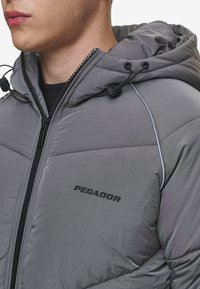 Pilkas izoliuotas džemperis su kapišonu, turintis priekinį užtrauktuką, reguliuojamus raiščius ir juodą prekės ženklą. Lygus tekstūra ir siuvinėjimas.
