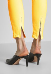 Gula smala jeans med blixtlåsdetailler vid anklarna. Kombinerat med svarta öppna klackar. Nærbild av underben och fotbeklädnad.