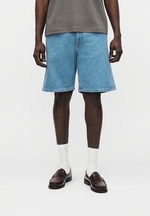 Moški, ki nosi svetlo modre jeans hlače, bele nogavice z elastičnim zgornjim robom, temno rjave usnjene loaferje in sivo majico, stoji pred enobarvnim ozadjem.