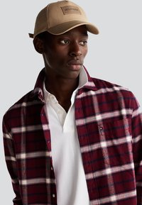 Cappello beige con una toppa logo rettangolare, camicia flanella a quadri rossi e blu navy sopra un polo bianco, dotata di bottoni e una tasca sul petto visibile.