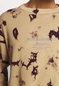 Suéter beige con patrones abstractos marrón oscuro, tejido de punto con textura, cuello redondo y texto impreso en azul en la parte delantera.