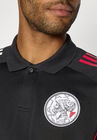 Sort polo-shirt med tre-knaps placket, der har et cirkulært Ajax-logo og røde striber på skuldrene. Fremstillet af struktureret stof.