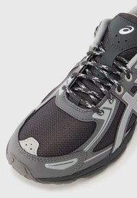 Γκρι αθλητικό παπούτσι με επάνω μέρος από δίχτυ, μαύρα κορδόνια και ανακλαστικές λεπτομέρειες. Διαθέτει ραμμένα ραφές και υφή στη λαστιχένια σόλα.