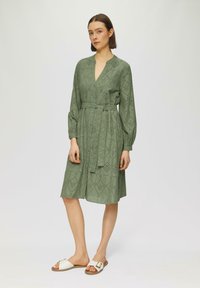 Abito verde, fino al ginocchio, con scollatura a V, maniche lunghe e vita legata. Presenta motivi ritagliati e bottoni blu. Indossato con sandali bianchi.