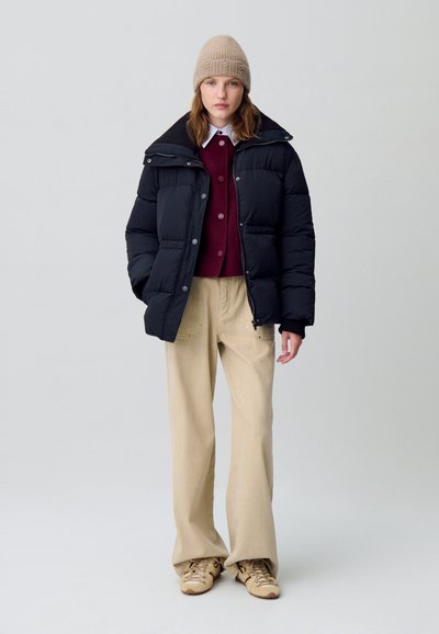 Giacca a vento nera con colletto, indossata sopra un maglione bordeaux e pantaloni beige. Include anche un berretto di maglia beige e sneakers color sabbia.