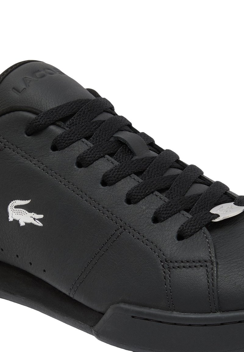 Baskets en cuir noir avec bout rond, surface texturée, lacets noirs, et un logo crocodile argenté sur le côté pour la marque.