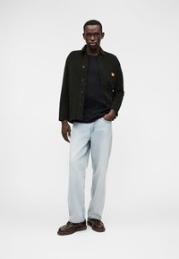 Jachetă din catifea de culoare verde închis cu nasturi, purtată peste o cămașă neagră. Blugi deschiși la culoare și pantofi maro completează ținuta.