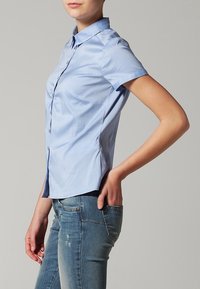 Chemise à manches courtes bleu clair en tissu lisse. Elle présente un col, une coupe ajustée et des coutures latérales, portée avec un jean déchiré.