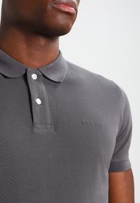 Närbild av en person som bär en grå Esprit-pikétröja med två vita knappar knäppta, vilket visar kragen och övre delen av torso.