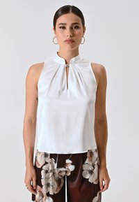 Haut sans manches blanc avec col haut et détail en trou de serrure, fabriqué à partir d'un tissu lisse. Doté d'un accent métallique décoratif au niveau du col.