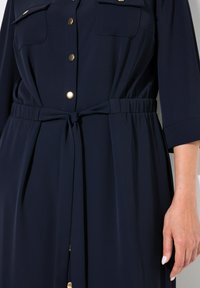Navyblaues Kleid mit tailliertem Schnitt, verziert mit goldenen Knöpfen und zwei Brusttaschen. Glatter Stoff und Dreiviertelärmel.