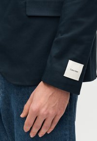 Το μανίκι του ναυτικού σακακιού φέρει μια λευκή ετικέτα με την επιγραφή "Calvin Klein." Διαθέτει ομαλή υφή υφάσματος και μινιμαλιστικό σχέδιο. Εμφανίζονται τζιν παντελόνια.