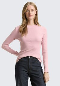 Long-sleeve pink ribbed top with a rounded neckline and frayed hem. Paired with dark blue jeans featuring a button and side pockets.