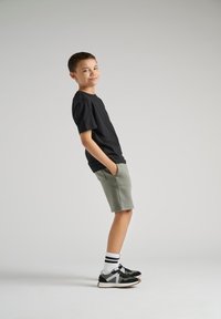 Sort bomulds T-shirt, lyse grønne shorts med lommer, sort og hvidt sportssko med striber, stående stilling mod en neutral baggrund.