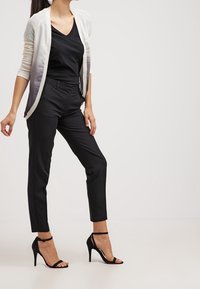 Kvinna iförd svart V-ringad topp, ombre cardigan, smala svarta byxor och svarta högklackade sandaler, stående mot en enkel ljus bakgrund.