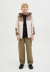 Giacca leggera a blocchi di colore in beige e marrone con cappuccio, zip frontale e polsini elasticizzati. Abbinata a una maglietta nera e pantaloni verdi.