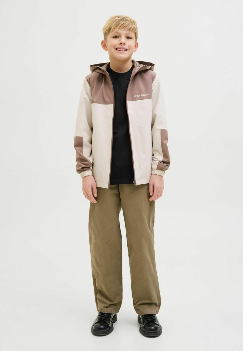 Giacca leggera a blocchi di colore in beige e marrone con cappuccio, zip frontale e polsini elasticizzati. Abbinata a una maglietta nera e pantaloni verdi.