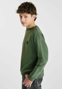 Groene katoenen sweatshirt met een ronde hals, lange mouwen en een klein zwart merkembleem op de borst, gecombineerd met donkere jeans.