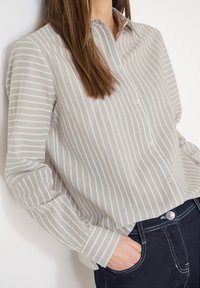 Femme portant une chemise à boutons à rayures verticales beige clair et blanche, rentrée dans un jean bleu foncé, la main dans la poche, sur fond blanc.