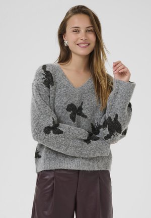 Lächelnde Frau mit silbernen Ohrringen, einem grauen, flauschigen Pullover mit schwarzen Schmetterlingsmustern und dunkelbraunen Lederhosen, die mit verschränkten Armen dasteht.