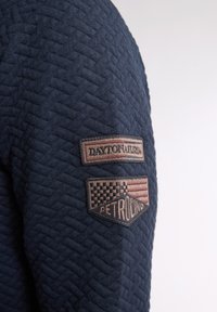 Pulover texturat de culoare bleumarin, cu țesătură în model de diamant. Are două insigne brodate pe mânecă, cu text și design în carouri.