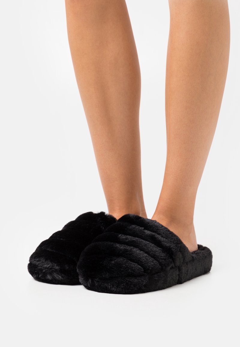 Chaussons noirs duveteux avec une texture douce et pelucheuse et un design matelassé, dotés d'un style à enfiler et d'un bout ouvert pour un port facile.