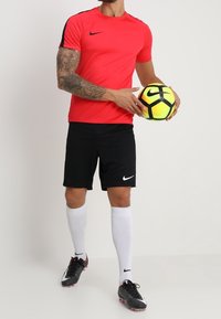 Punainen urheilupaita mustilla yksityiskohdilla, mustat shortsit ja valkoiset polvisukat. Pelaaja pitää mustaa ja keltaista jalkapalloa käsissään.