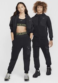Schwarze Nike Pro Sportbekleidung, bestehend aus einem kurzen Top und einer Jacke mit Reißverschluss, kombiniert mit Jogginghosen mit Taschen und schwarzen Turnschuhen.