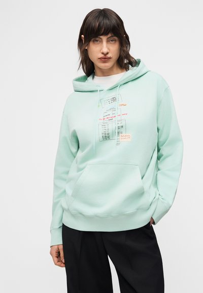 Άτομο που φοράει φούτερ με κουκούλα σε μέντα πράσινο χρώμα με σχέδια ημερολογίου και χειρόγραφου κειμένου, συνδυασμένο με χαλαρό μαύρο παντελόνι, σε γκρι φόντο.