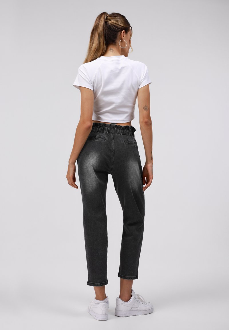 Femme avec queue de cheval portant un t-shirt blanc court, un jean noir délavé taille haute, des baskets blanches, debout de dos sur un fond uni.