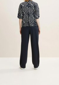 Person steht mit dem Rücken zur Kamera, trägt schwarze weite Hosen, weiße Schuhe und ein schwarzes kurzärmliges Hemd mit weißen Paisley-Mustern.