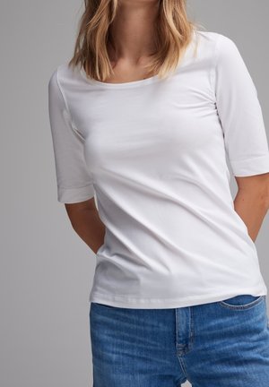 Vrouw draagt een effen wit shirt met korte mouwen en blauwe spijkerbroek, staat met haar handen achter haar rug tegen een grijze achtergrond.