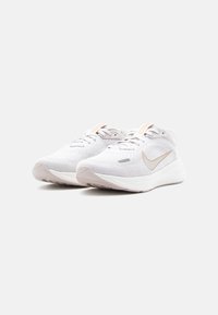 Ζευγάρι λευκά πλεκτά αθλητικά παπούτσια Nike με μπεζ λογότυπα swoosh και ανοιχτό γκρι σόλες σε απλό φόντο.