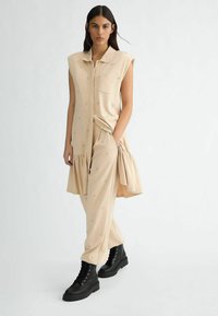 Beiges ärmelloses Outfit mit einer Tasche, rüschigem Saum und gestickten Akzenten; kombiniert mit schwarzen, geschnürten Stiefeletten und einer glatten Textur.