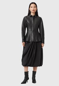 Camicia in pelle nera con colletto, corpo aderente e chiusura a bottoni, abbinata a una gonna nera voluminosa e drappeggiata e stivaletti.