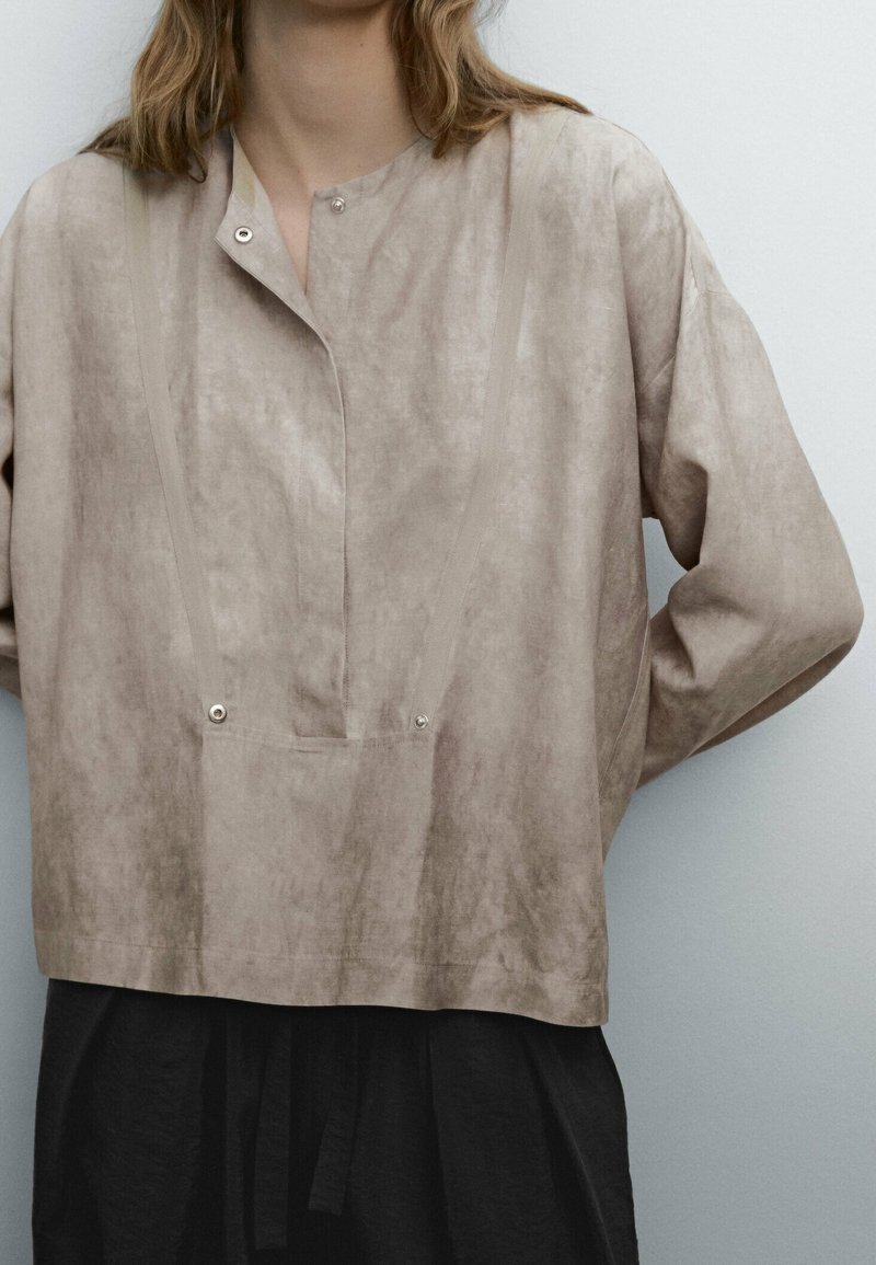 Haut beige clair à manches longues avec une coupe décontractée, doté d'une fermeture à boutons-pression et de détails de coutures verticales. Tissu texturé avec un léger éclat.