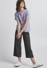 Ljuslila t-shirt med "BEYOND TIME" i röd tryck, i kombination med mörkgrå cropped byxor och vita sneakers. Slitstark tyg, avslappnad passform.