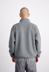 Grå fleecetröja med hög krage, texturerad utsida och elastisk fåll. Har en avslappnad passform och långa ärmar. Bakifrån sedd.