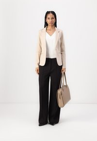 Blazer aderente beige, top bianco a scollo a V, pantaloni neri a gamba larga e borsa color taupe. Tessuti lisci, design su misura e colori neutri.