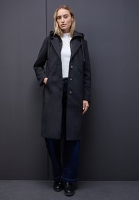 Zwarte lange jas met capuchon, dubbelzijdige knoopsluiting, zijzakken en een gladde textuur, gedragen over een witte top en donkerdenim.