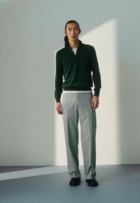 Groene ritstrui, witte shirt eronder, grijze geplooide broek en zwarte schoenen. De trui heeft een hoge kraag en geribde manchet.