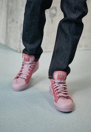 Person iført mørke jeans og pinke høje sneakers med snørebånd går på en lyseblå plettet gulv.